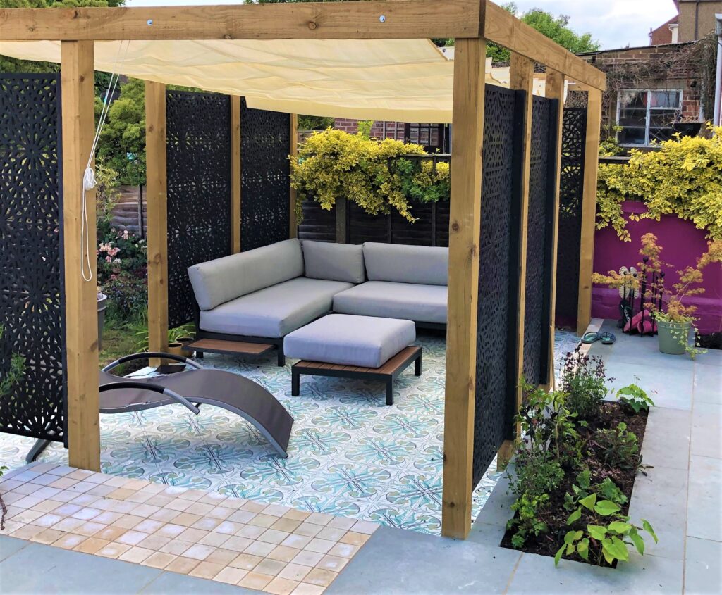 Wooden-framed pergola with black metal panels cut with islamic patterns. Within the space is low level seating with grey covers, all in a hard-landscaped garden.