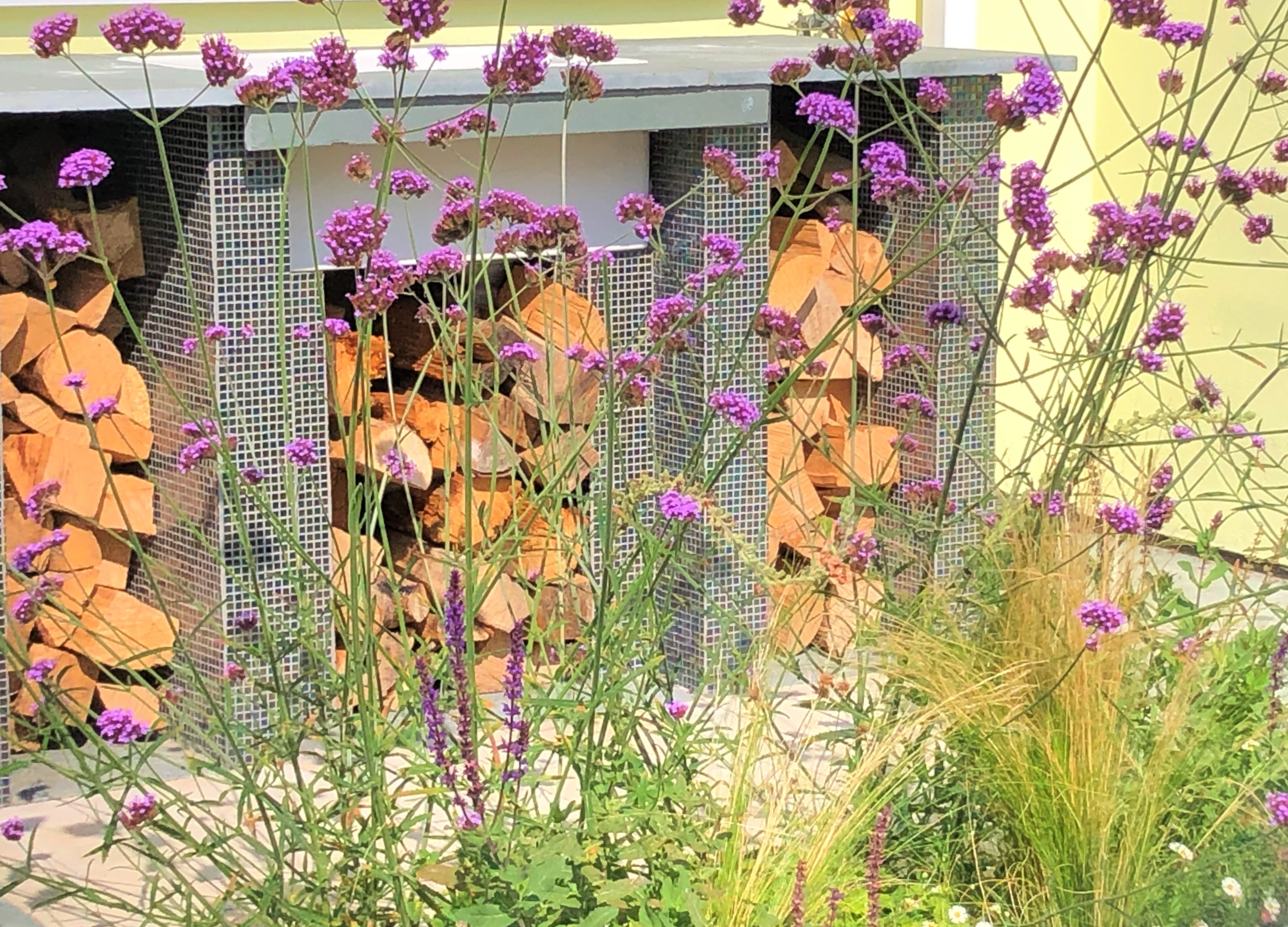 An outdoor sink area, with added wood storage, clad in mosaic tiles is seen through colourful planting that springs from within the patio
