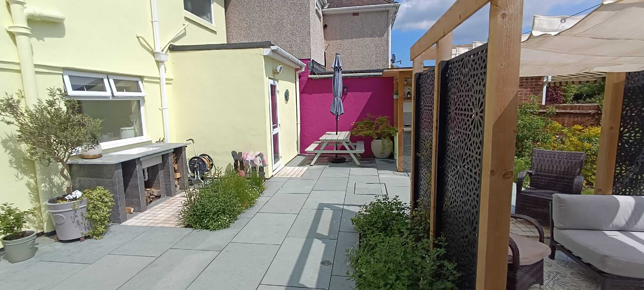 A patio with an outdoor sink and woodstore, a bar and a seating area with lush planting and colour all around